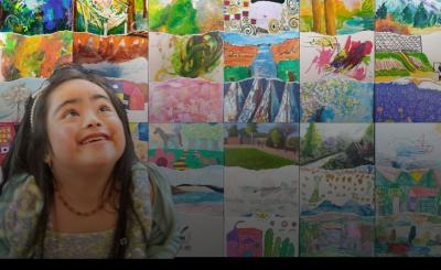 A little girl looks up smiling. Behind is her are different paintings.
