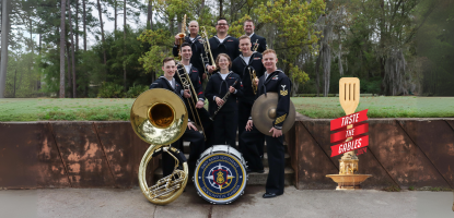 5 musicians stand in Navy uniform holding their instruments in front of a wall with grass and forest