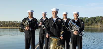 Five people in Navy uniforms hold brass instruments and stand on a bridge on a lake