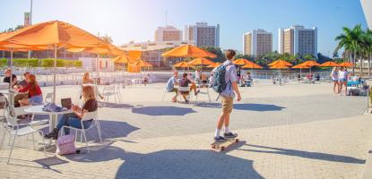 Beautiful sunny day and blue sky, college students sit under orange umbrellas while a man skateboards past the plaza