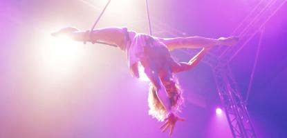 Strongly backlit by purple lights, an acrobat does a split in the air on ropes