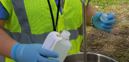 Official in lime green vest, blue gloves pours water into a container from a temporary sink out outside by grass