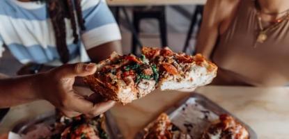 A slice of square pizza is raised into focus with a man and a woman smiling in the background