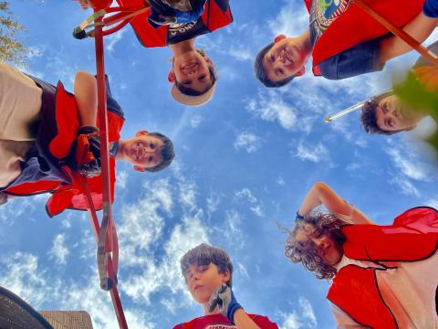 Point of view the ground looking up, kids in orange vests with tools look down. Behind them a bright blue sky freckled with white clouds