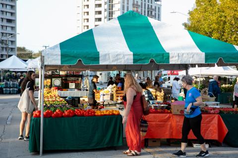 Two women walk by a green and white tent covering fruits and vegetables