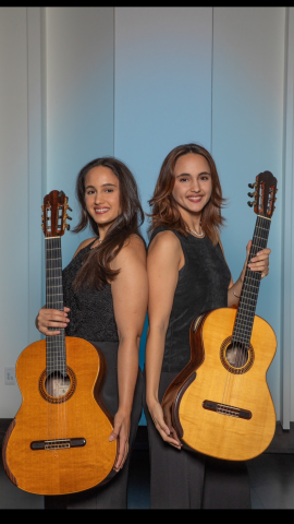 Inside with white background, two women wearing sleeveless black clothes stand back to back holding guitars