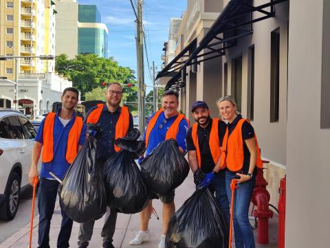 5 adults in orange vests carry trash bags and smile at the camera on a beautiful, shaded sidewalk on sunny day