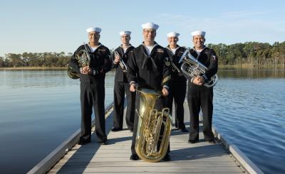 Five people in Navy uniforms hold brass instruments and stand on a bridge on a lake