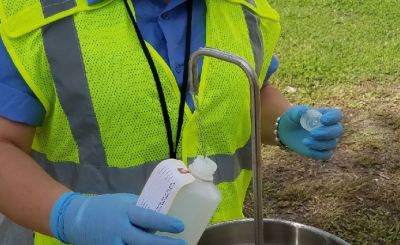 Official in lime green vest, blue gloves pours water into a container from a temporary sink out outside by grass