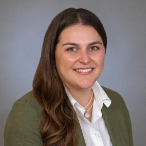 Anna Pernas smiles in a green jacket, white shirt in front of a gray background