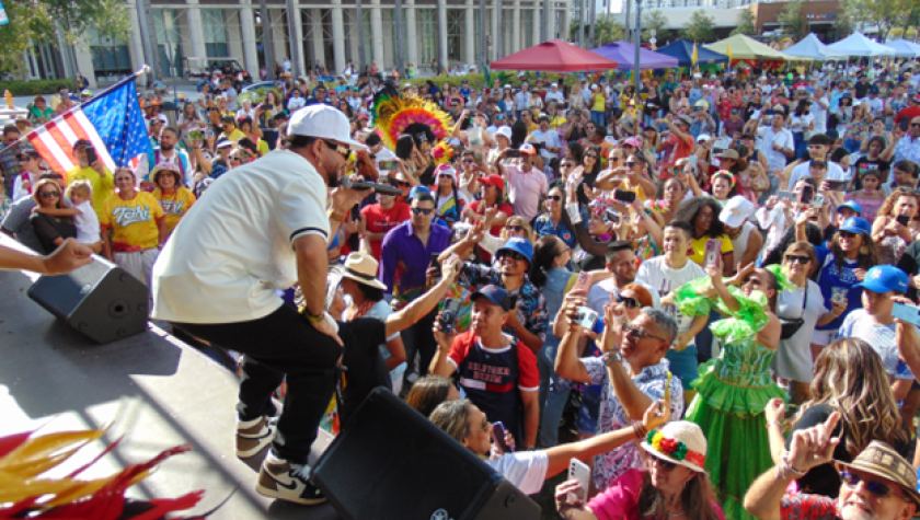 A man on stage sings into a microphone to a large, packed crowd of people on a hot, sunny day