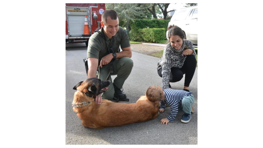 An K9 officer in green kneels besides a mother with her baby petting a German shepherd dog